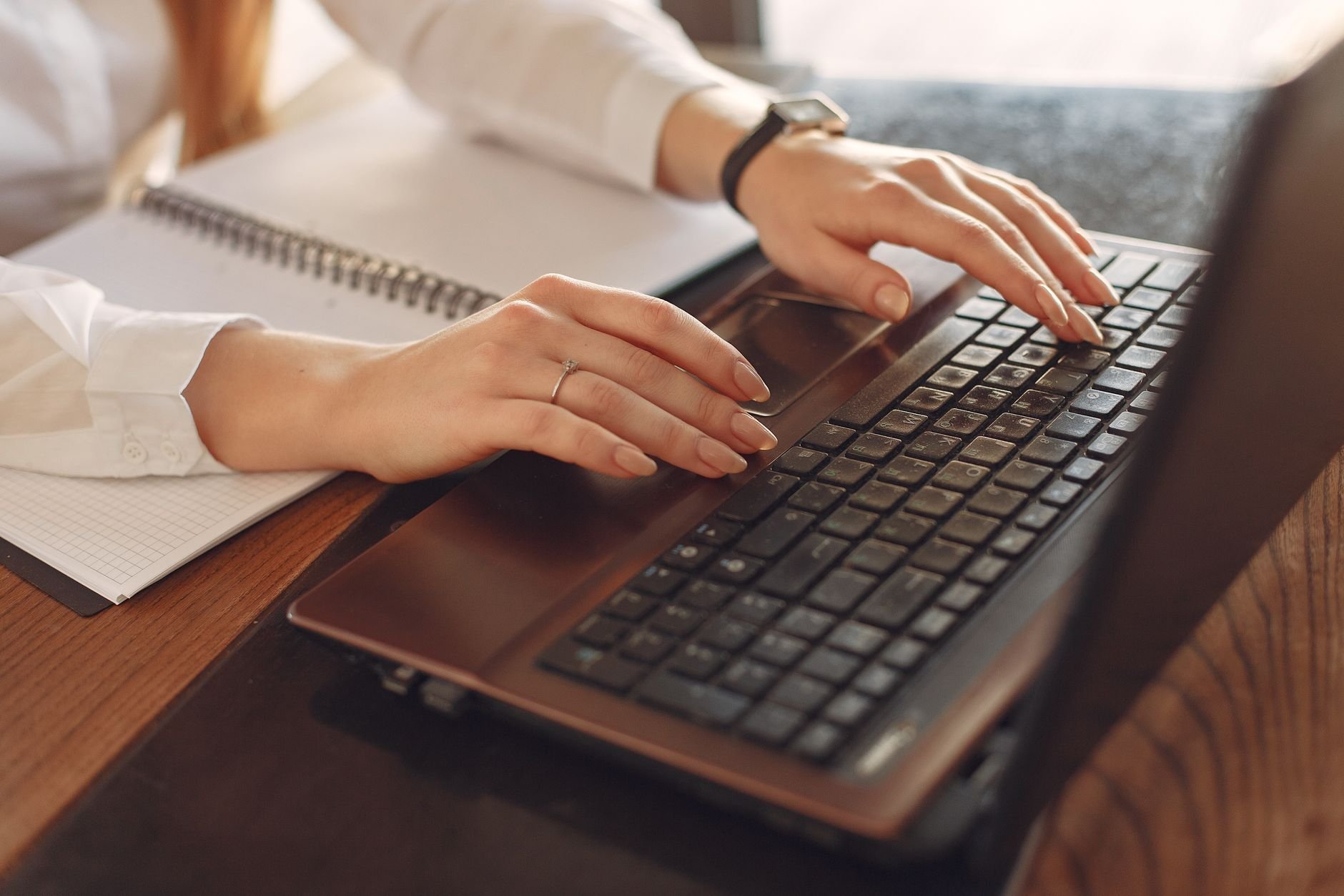 Close-up of a person typing on a laptop with a notebook nearby